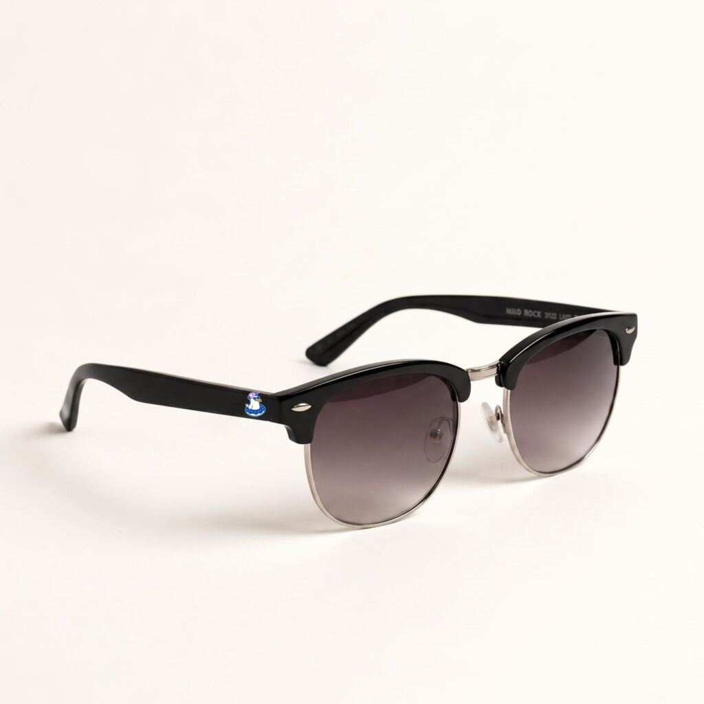Lunettes de soleil homme tendance de la collection été Marseille par Chouchou Beignet. Le modèle présente une monture de forme Clubmaster en acétate noir profond sur la partie supérieure et en métal argenté sur la partie inférieure. Les verres affichent une teinte dégradée allant du gris foncé au transparent. Sur la branche en plastique noir, on aperçoit le logo Chouchou Beignet représentant un oiseau avec une bouée. L'accessoire plage est photographié de trois-quarts sur un fond blanc uni et épuré.