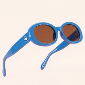 Lunettes de soleil au style rétro et tendance, arborant une monture ovale et galbée en acétate brillant de couleur bleu azur. Les verres teintés d'un marron chaud offrent une protection douce. Présentée en lévitation de trois-quarts face sur un fond beige rosé très épuré, cette paire met en valeur le logo Chouchou Beignet avec son gabian en bouée sur la branche droite, l'accessoire plage idéal pour la collection été Marseille.