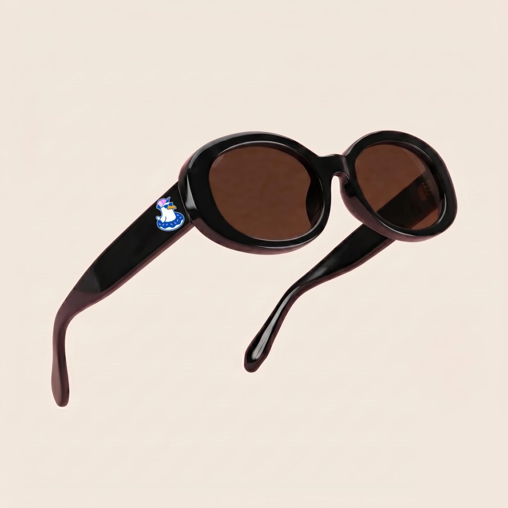 Lunettes de soleil au style rétro et tendance, arborant une monture ovale et galbée en acétate brillant de couleur noir profond. Les verres teintés d'un marron chaud offrent une protection douce. Présentée en lévitation de trois-quarts face sur un fond beige rosé très épuré, cette paire met en valeur le logo Chouchou Beignet avec son gabian en bouée sur la branche droite, l'accessoire plage idéal pour la collection été Marseille.