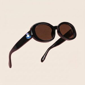 Lunettes de soleil au style rétro et tendance, arborant une monture ovale et galbée en acétate brillant de couleur noir profond. Les verres teintés d'un marron chaud offrent une protection douce. Présentée en lévitation de trois-quarts face sur un fond beige rosé très épuré, cette paire met en valeur le logo Chouchou Beignet avec son gabian en bouée sur la branche droite, l'accessoire plage idéal pour la collection été Marseille.
