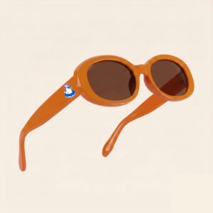 Lunettes de soleil au style rétro et tendance, arborant une monture ovale et galbée en acétate brillant de couleur orange vif. Les verres teintés d'un marron chaud offrent une protection douce. Présentée en lévitation de trois-quarts face sur un fond beige rosé très épuré, cette paire met en valeur le logo Chouchou Beignet avec son gabian en bouée sur la branche droite, l'accessoire plage idéal pour la collection été Marseille.