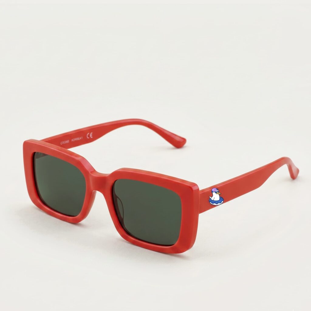 Lunettes de soleil femme tendance avec monture en plastique rouge intense et forme carrée, verres gris foncé, posées sur fond clair minimaliste, logo Chouchou Beignet sur la branche, accessoire plage de la collection été Marseille.