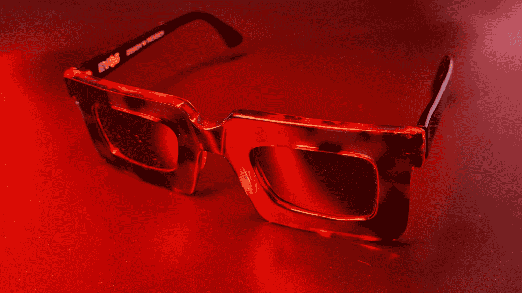 Gros plan d'une paire de lunettes de soleil carrées et épaisses, d'une couleur rouge intense et brillante, posée sur une surface réfléchissante.