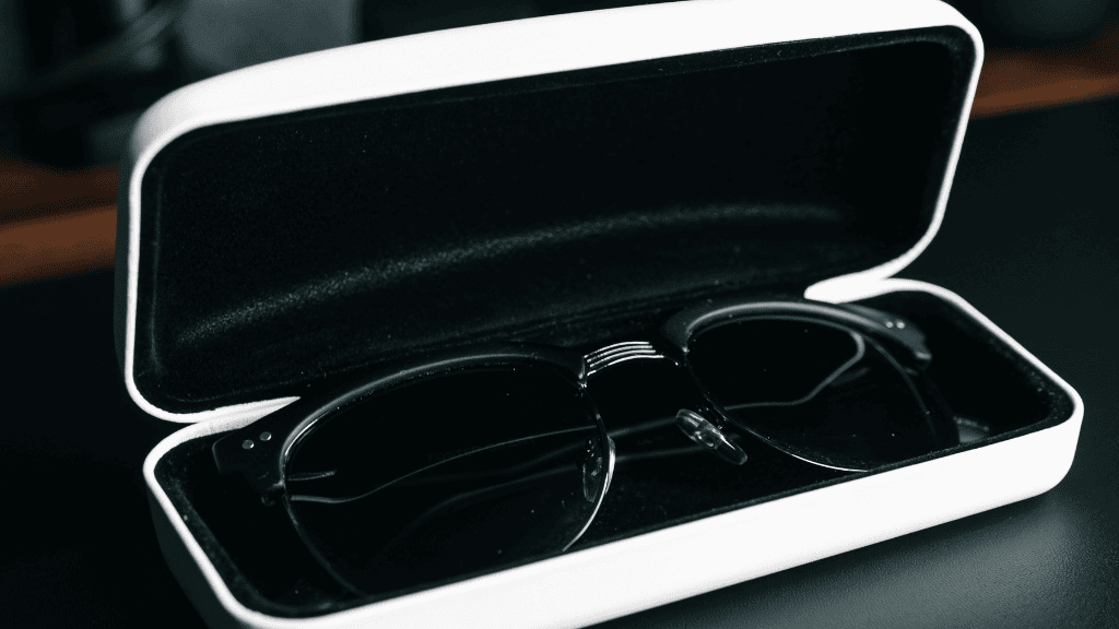Lunettes de soleil noires protégées dans un étui rigide blanc avec intérieur en velours noir, posé sur une surface sombre.
