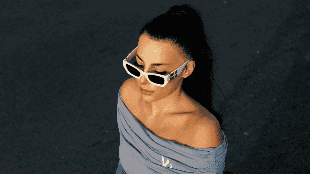 Jeune femme en tenue de sport bleue portant des lunettes de soleil de luxe à monture blanche épaisse, tenant un café glacé.