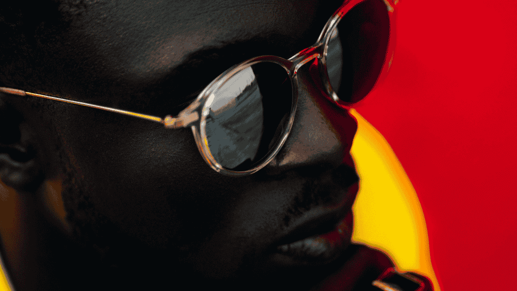 Portrait d'un homme portant des lunettes de soleil tendance avec une monture transparente et des verres sombres, sur un fond contrasté rouge et jaune.