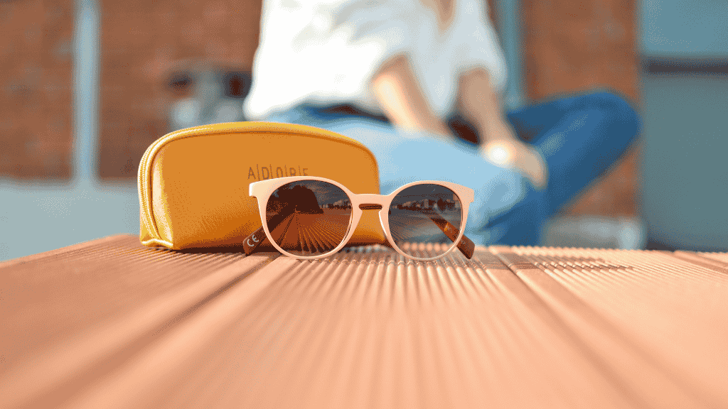 Paire de lunettes de soleil rondes rose pâle posée sur un deck en bois à côté d'un étui jaune moutarde, avec une personne floutée en arrière-plan.