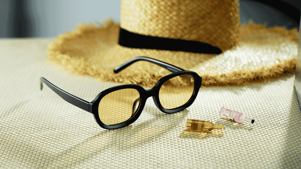 Paire de lunettes de soleil aux formes arrondies et monture noire posée sur une nappe tressée devant un chapeau de paille.
