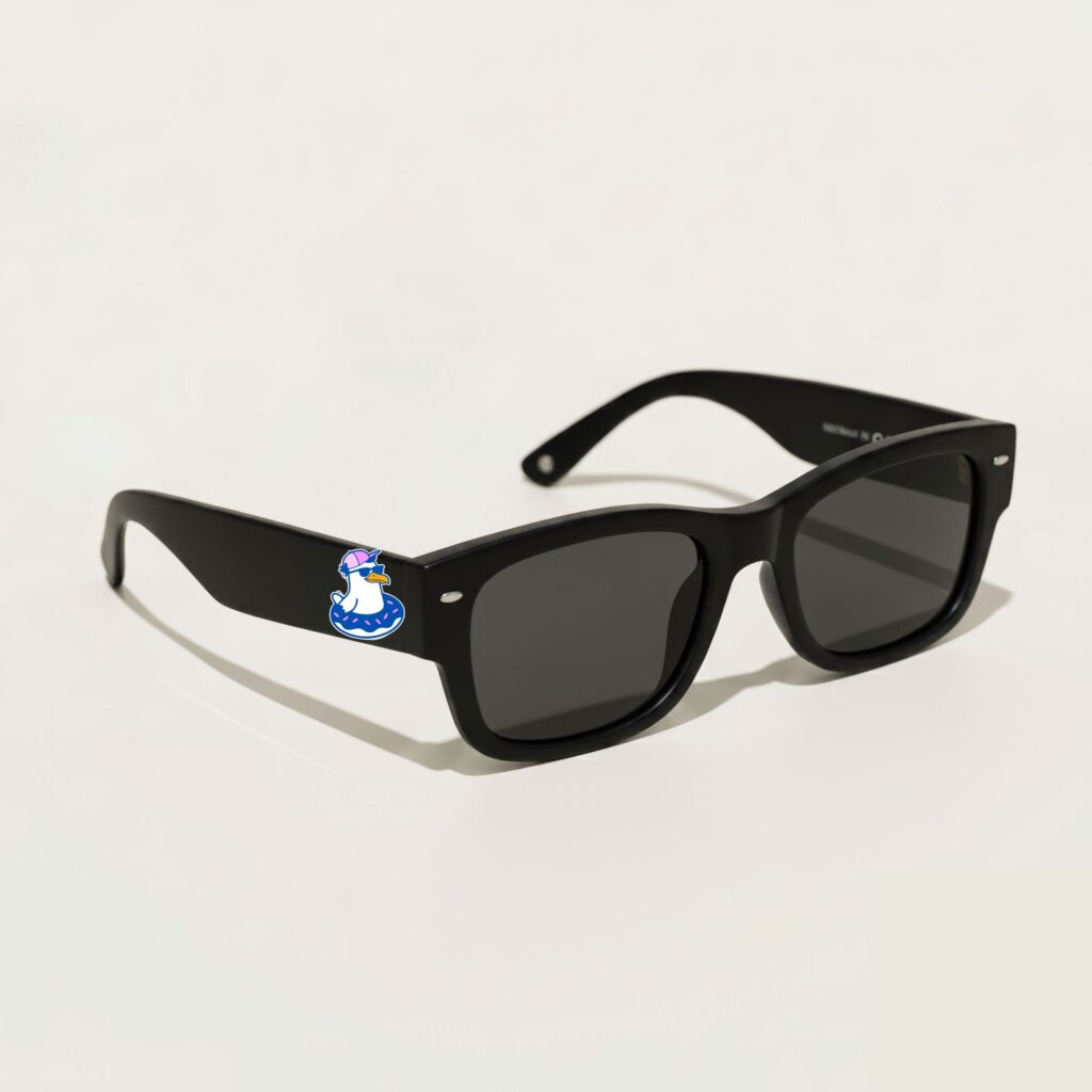 Lunettes de soleil femme au style Wayfarer tendance, présentant une monture épaisse en acétate noir profond au fini mat. Les verres de teinte gris foncé s'accordent parfaitement avec le style sobre de la paire. Photographié de trois-quarts face sur un fond blanc crème épuré, cet accessoire plage indispensable de la collection été Marseille affiche fièrement sur sa branche gauche le logo Chouchou Beignet avec son gabian à casquette dans sa bouée.