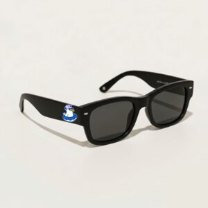 Lunettes de soleil femme au style Wayfarer tendance, présentant une monture épaisse en acétate noir profond au fini mat. Les verres de teinte gris foncé s'accordent parfaitement avec le style sobre de la paire. Photographié de trois-quarts face sur un fond blanc crème épuré, cet accessoire plage indispensable de la collection été Marseille affiche fièrement sur sa branche gauche le logo Chouchou Beignet avec son gabian à casquette dans sa bouée.