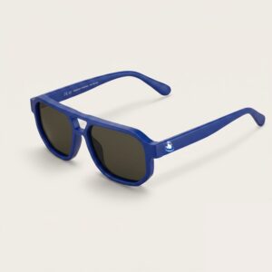 Lunettes de soleil femme tendance avec monture en métal bleu azur et forme aviateur, verres gris foncé, posées sur fond clair minimaliste, logo Chouchou Beignet sur la branche, accessoire plage de la collection été Marseille.