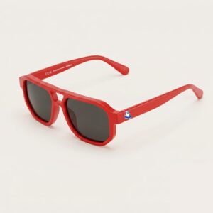 Lunettes de soleil femme tendance avec monture en métal rouge intense et forme aviateur, verres gris foncé, posées sur fond clair minimaliste, logo Chouchou Beignet sur la branche, accessoire plage de la collection été Marseille.