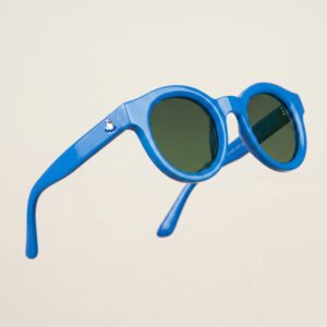 Lunettes de soleil femme tendance avec monture en plastique bleu azur et forme ronde, verres gris foncé, posées sur fond clair minimaliste, logo Chouchou Beignet sur la branche, accessoire plage de la collection été Marseille.