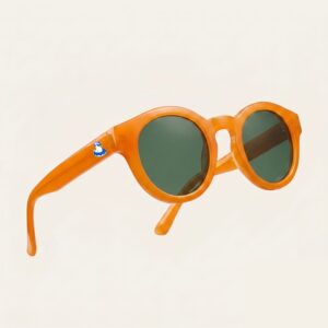 Lunettes de soleil femme tendance avec monture en plastique orange vif et forme ronde, verres gris foncé, posées sur fond clair minimaliste, logo Chouchou Beignet sur la branche, accessoire plage de la collection été Marseille.