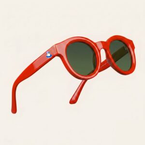 Lunettes de soleil femme tendance avec monture en plastique rouge intense et forme ronde, verres gris foncé, posées sur fond clair minimaliste, logo Chouchou Beignet sur la branche, accessoire plage de la collection été Marseille.
