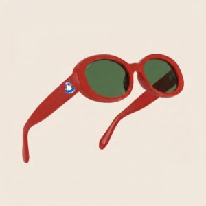 Lunettes de soleil femme tendance avec monture en plastique rouge intense et forme ovale, verres verts foncés, posées sur fond clair minimaliste, logo Chouchou Beignet sur la branche, accessoire plage de la collection été Marseille.