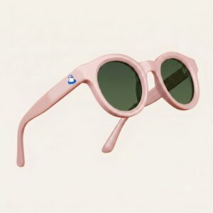 Lunettes de soleil au style rétro-chic et tendance, arborant une monture de forme ronde en acétate brillant de couleur rose bonbon. Les verres foncés affichent une teinte gris-vert hautement protective. Présentée de trois-quarts face en lévitation sur un fond beige rosé uni et épuré, cette paire met en valeur le logo Chouchou Beignet avec son gabian en bouée sur la branche droite, l'accessoire plage incontournable de la collection été Marseille.