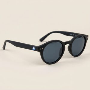 Lunettes de soleil femme au style Pantos tendance, se distinguant par une monture arrondie en acétate mat de couleur noir profond. Les verres foncés affichent une teinte gris-noir hautement protectrice. Présenté de trois-quarts face sur un fond beige sable très doux et épuré, cet accessoire plage incontournable met en avant le logo Chouchou Beignet avec son gabian en bouée sur la branche droite, une pièce maîtresse pour la collection été Marseille.