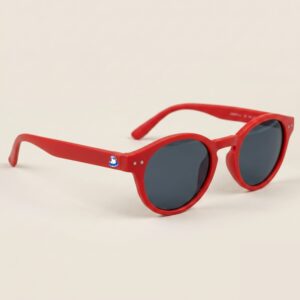 Lunettes de soleil femme au style Pantos tendance, caractérisées par une monture arrondie en acétate mat de couleur rouge intense. Les verres foncés affichent une teinte gris-bleu très protectrice. Présenté de trois-quarts face sur un fond beige sable très doux et épuré, cet accessoire plage indispensable met en avant le logo Chouchou Beignet avec son gabian en bouée sur la branche droite, idéal pour compléter la collection été Marseille.