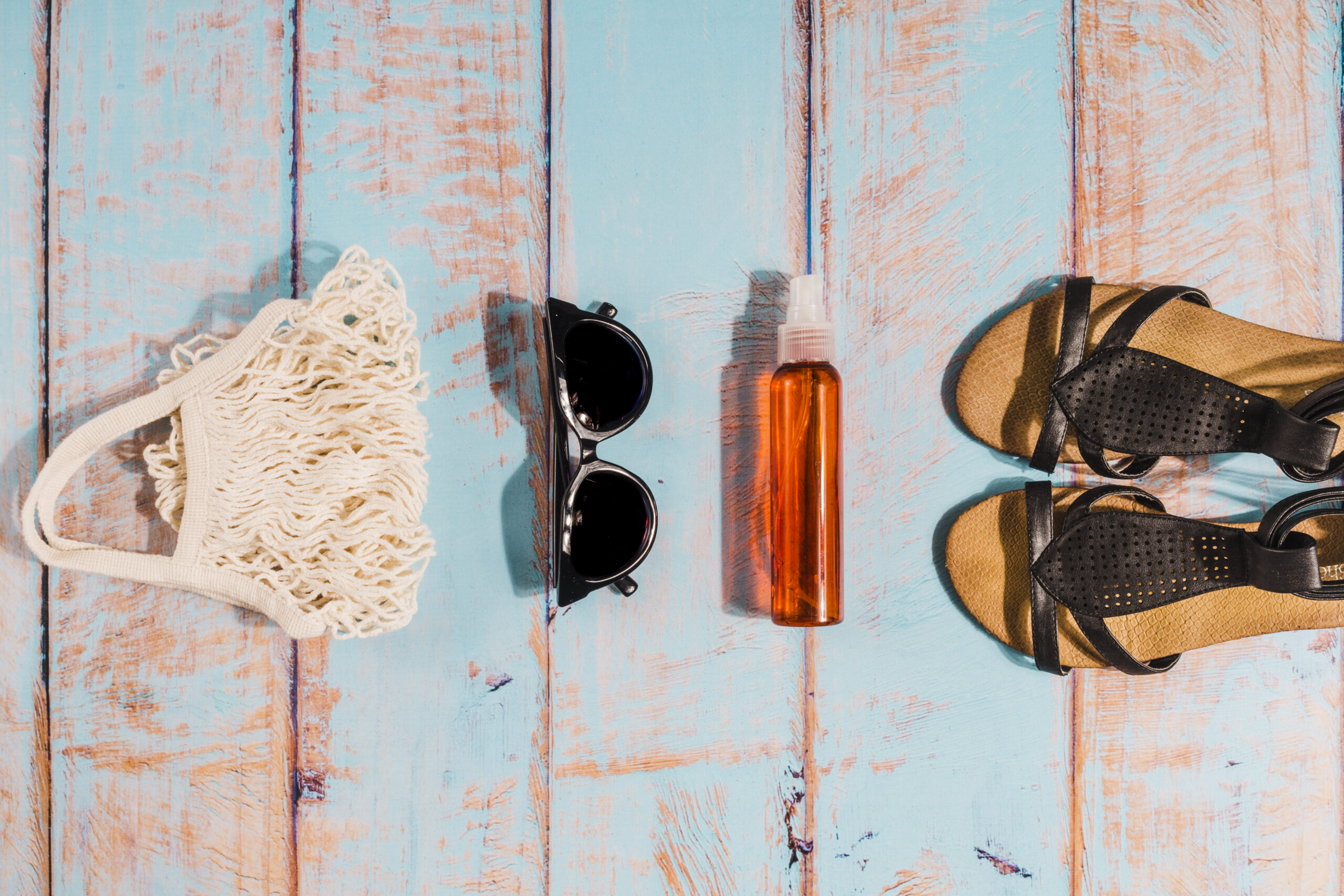 Vue de dessus d'accessoires de plage (sac filet, lunettes de soleil, spray solaire et sandales) alignés sur une table en bois bleu vieilli.